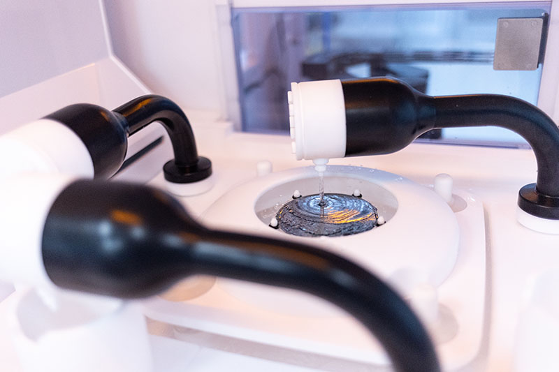 stone-factory-05 A close-up of a single-wafer wet etching process for Indium Phosphide (InP), showing a precision nozzle dispensing a chemical etchant onto the center of a spinning wafer within an automated JST Manufacturing semiconductor tool.